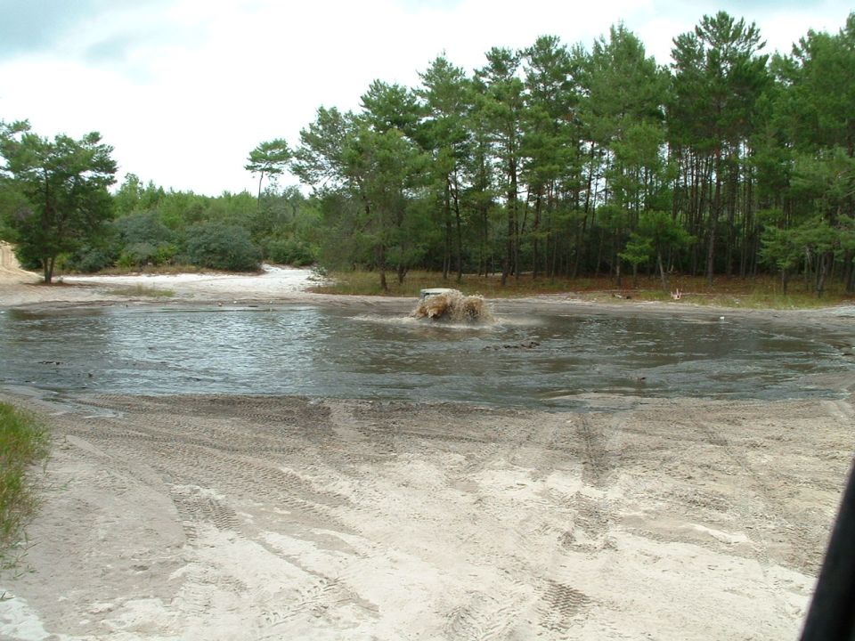 Off-road vehicle driving through a shallow body of water with trees in the background. Tire tracks are visible in the sand