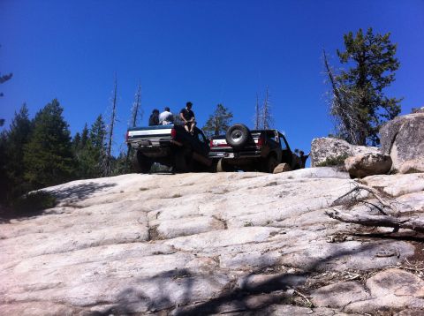 1990 Ford Bronco - Two SUVs navigate a rocky trail under a clear blue sky, with people riding in the back of the lead vehicle.