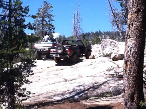 1990 Ford Bronco - Two SUVs navigate a challenging off-road path with large rocks and trees under a clear blue sky.