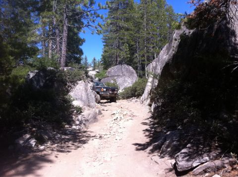 1990 Ford Bronco - A dark SUV navigates a challenging, rocky trail surrounded by dense foliage and large rock formations.