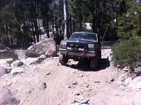 1990 Ford Bronco - A dark-colored Ford Bronco navigates a rocky, off-road trail surrounded by trees.