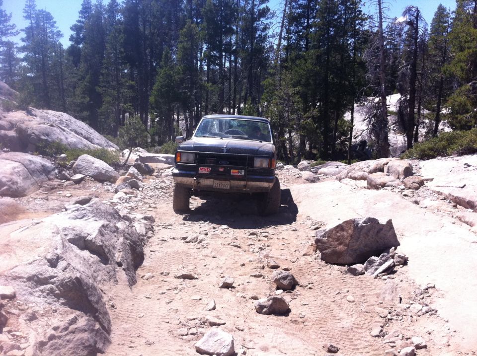 1987 Ford Bronco navigating a rocky off-road trail with trees in the background