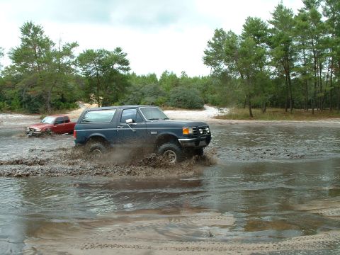 1995 Ford Bronco - Two SUVs navigate a muddy water crossing on a dirt road with trees lining the area.