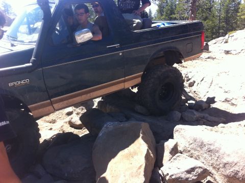 1990 Ford Bronco - A dark green Ford Bronco with a tan stripe is shown navigating a rocky trail, with the driver visible behind the wheel.