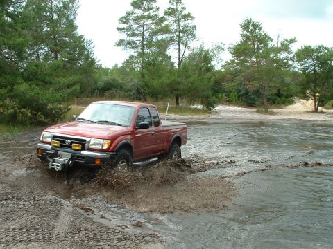 1995 Ford Bronco - A red Toyota Tacoma navigates through a muddy off-road area, kicking up water and dirt.