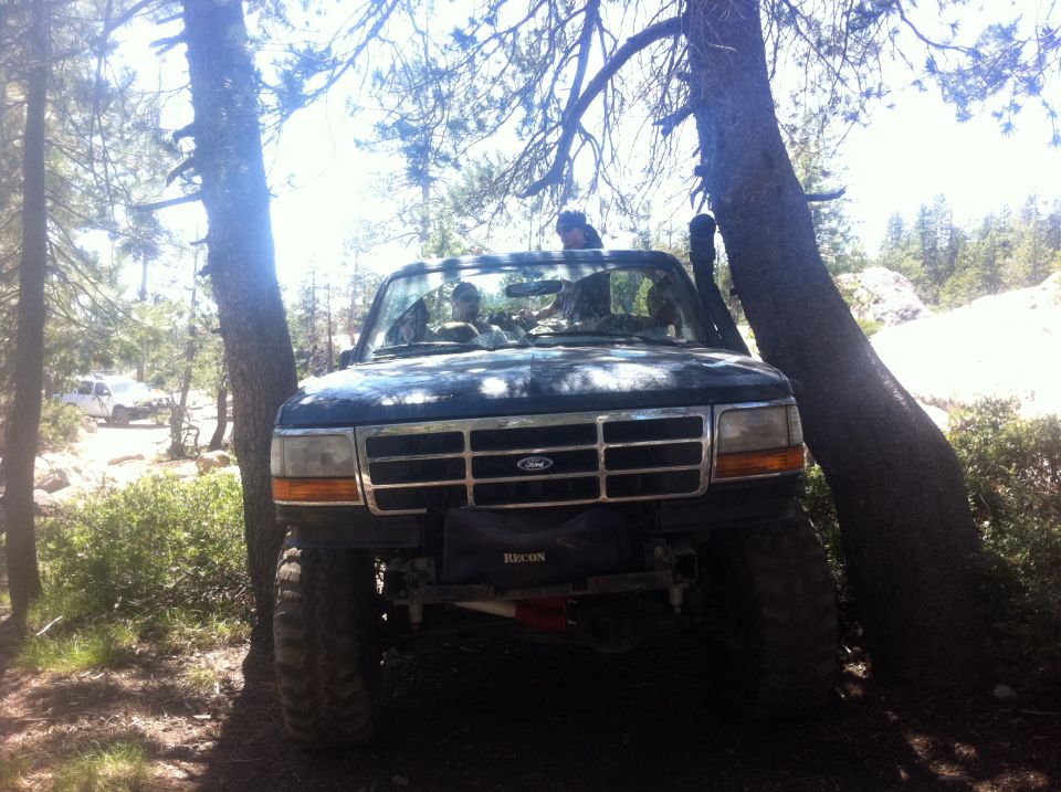 Front view of a dark-colored Ford Bronco with large tires, parked between trees on a dirt trail. Another white SUV is visible in the background