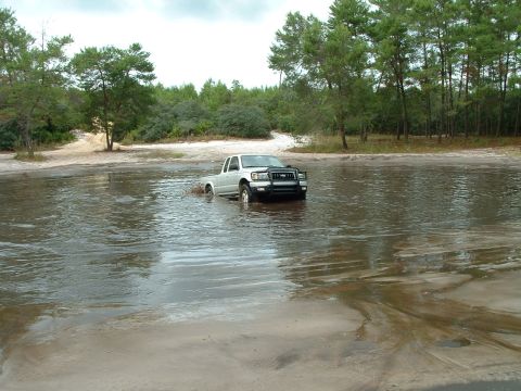 1995 Ford Bronco - Pickup truck navigates through a water crossing on a dirt road surrounded by trees.