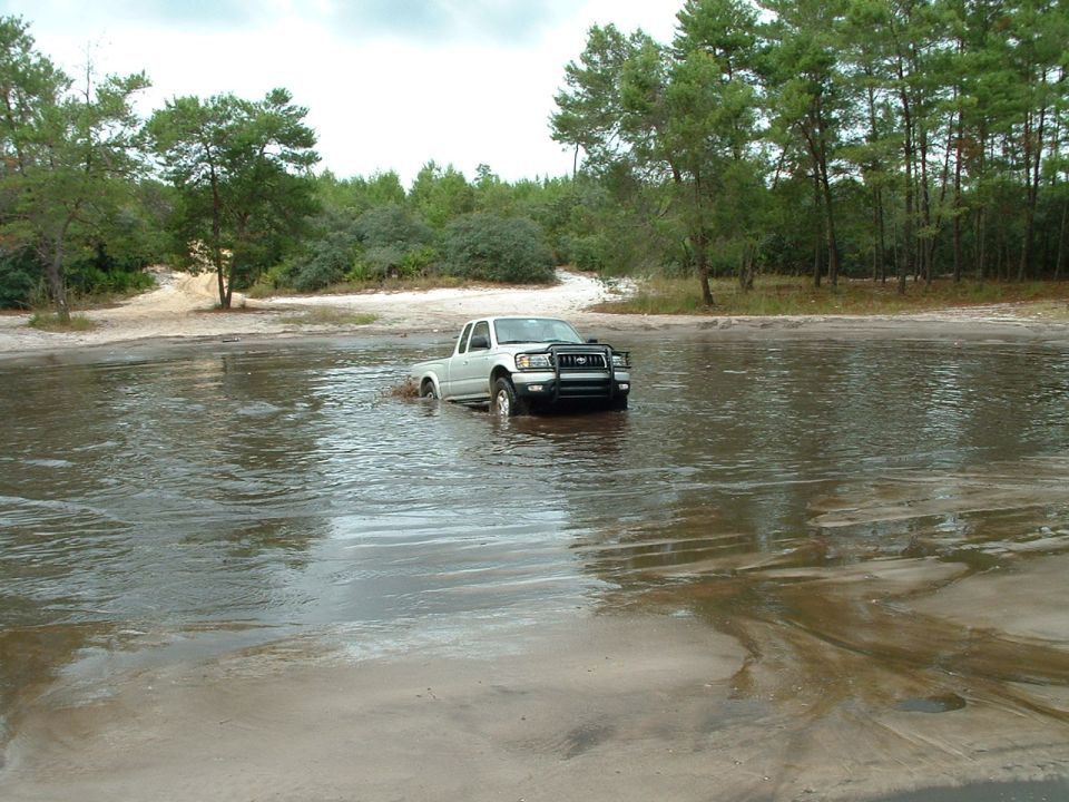 Silver pickup truck with black bull bar driving through shallow water and mud