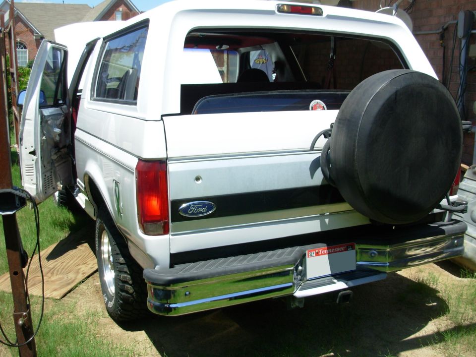 Rear view of white Ford Bronco with black spare tire cover, chrome bumper, and Tennessee plate, driver's door open