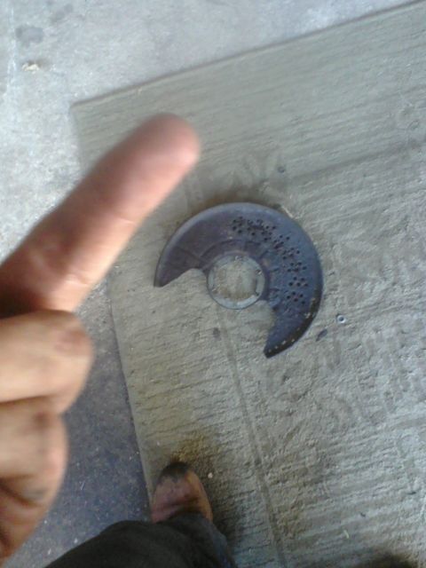 1988 Ford Bronco - A brake dust shield lies on a concrete surface, with a finger pointing towards it.
