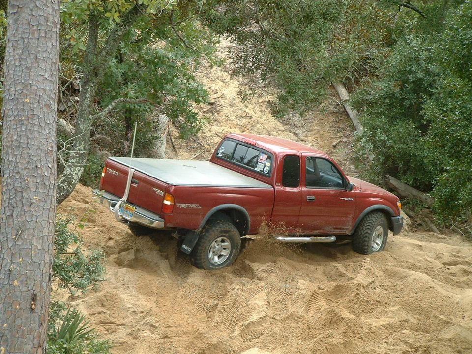 Red Toyota Tacoma TRD off-road pickup truck climbing a sand dune. Visible badges include Toyota and TRD