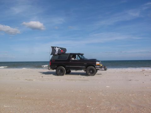 1995 Ford Bronco - A black SUV is parked on the beach with the ocean in the background under a blue sky.