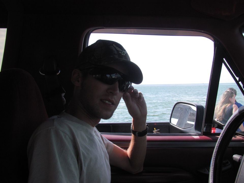 Guy in a baseball cap and sunglasses adjusts his shades inside a truck cab, looking out at the ocean. Another person is visible in the passenger seat