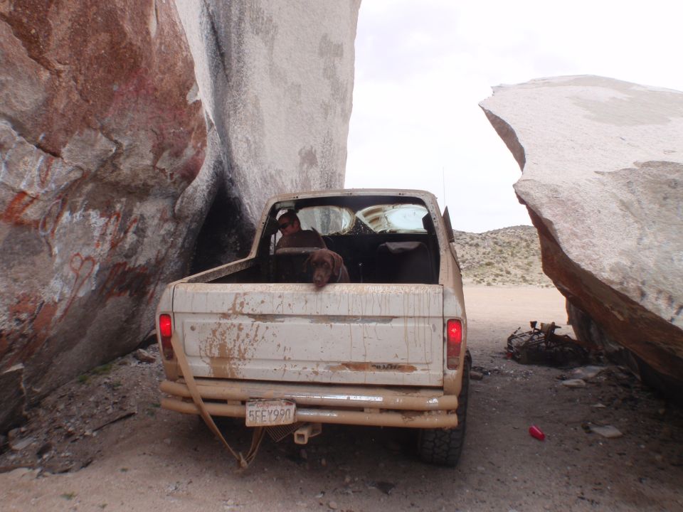 Mud-splattered beige Ford pickup truck stuck between large rocks, rear view