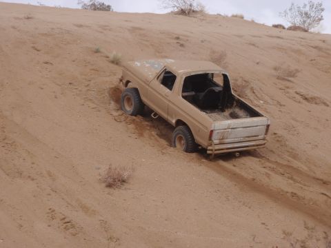 1978 Ford Bronco - A tan 1978 Ford Bronco is partially submerged in sand on a dune, with its rear facing the camera.