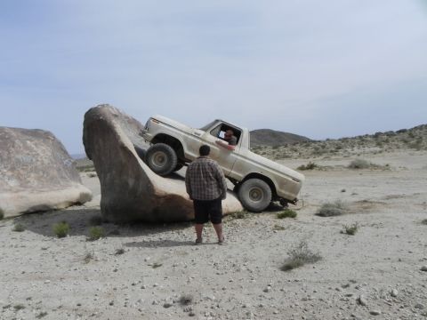 1978 Ford Bronco - A dusty white topless full-size Ford Bronco climbs nose-up onto a large sloped desert boulder, front wheels on the rock while a spotter in…