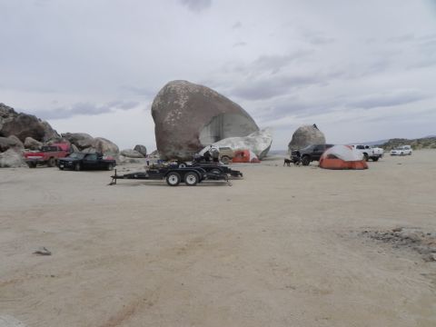 1978 Ford Bronco - Desert camp at Giant Rock, with an empty black tandem-axle car hauler trailer parked on the sand in the foreground. A red older pickup, a d…