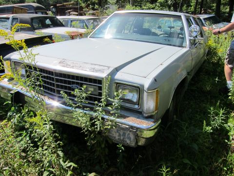 1991 Ford LTD Crown Victoria - A light blue 1991 Ford Crown Victoria LTD sits in a junkyard, partially obscured by weeds and other vehicles.