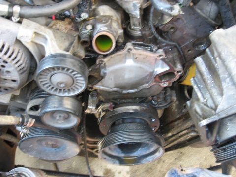 1994 Ford Bronco - Front of an engine with the serpentine belt and accessories partly stripped, exposing the cast water pump housing, the ribbed crank pulley…