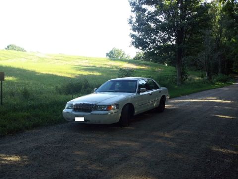 2000 Mercury Grand Marquis - A white 2000 Mercury Grand Marquis sedan is parked on a gravel road beside a grassy embankment under a tree.