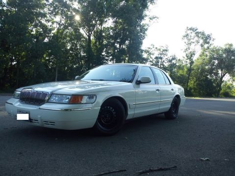 2000 Mercury Grand Marquis - A white 2000 Mercury Grand Marquis is seen from a three-quarter front view, parked outdoors with trees and a bright sky.