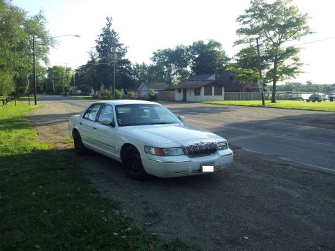 2000 Mercury Grand Marquis - A white 2000 Mercury Grand Marquis is parked on the side of a road with houses and trees in the background.