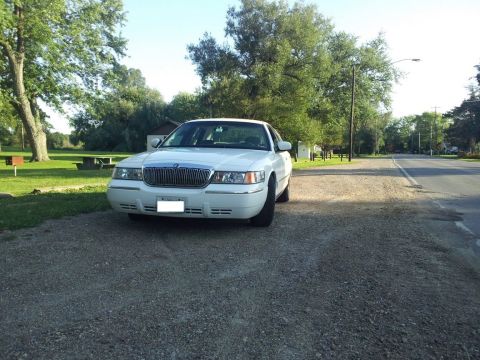 2000 Mercury Grand Marquis - A white 2000 Mercury Grand Marquis is parked on a gravel surface next to a road with trees in the background.