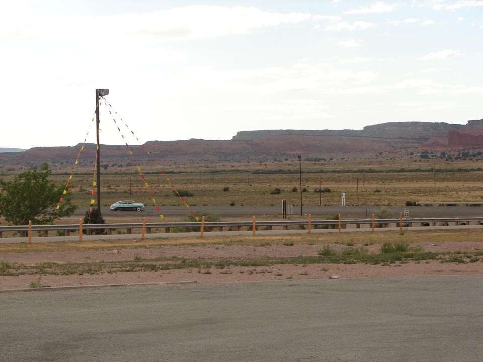 White sedan driving on highway, desert landscape with mesas behind. Pole with flags in foreground