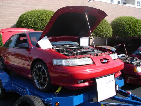 1992 Ford Taurus - A red sedan is displayed on a trailer with its hood open, revealing the engine compartment. Another vehicle is visible behind it.