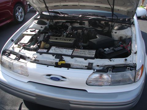 1992 Ford Taurus - The engine bay of a white 1992 Ford Taurus is displayed with the hood open, revealing the 3.8L V6 Fuel Injected engine.