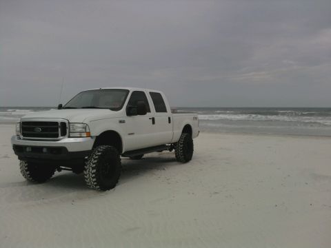 1995 Ford Bronco - A lifted white Ford F-250 pickup truck sits on a sandy beach with the ocean in the background under an overcast sky.