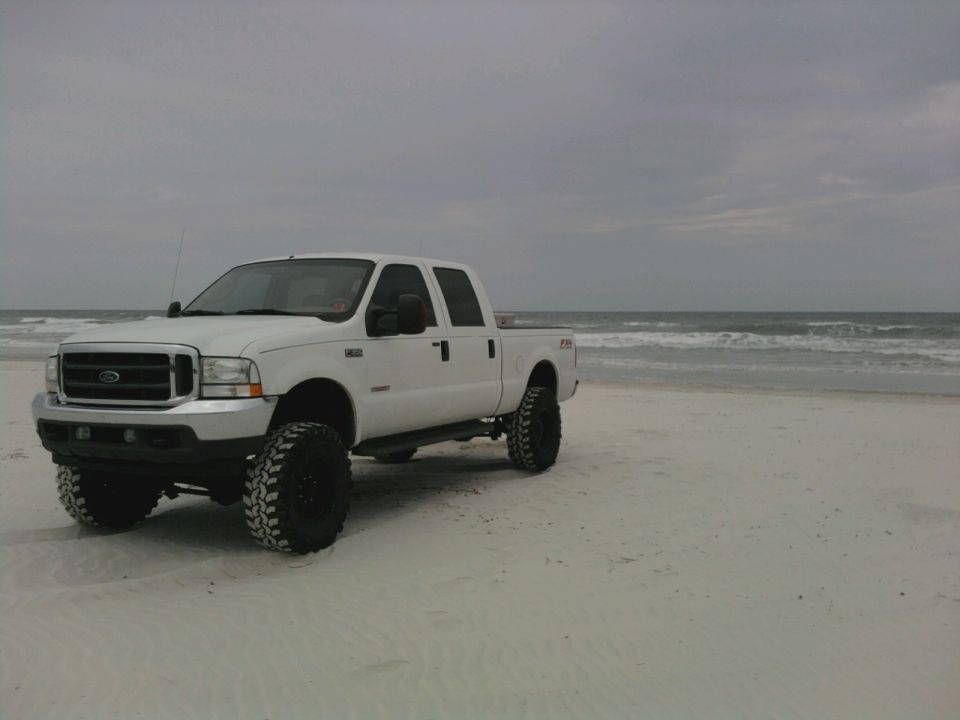 White Ford F-250 pickup truck with lifted suspension and large tires parked on a sandy beach near the ocean