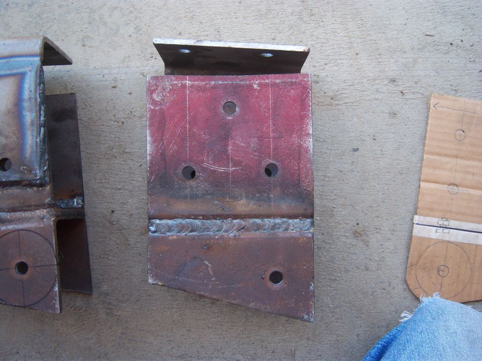 Two metal brackets with holes drilled, one is red. Cardboard template nearby