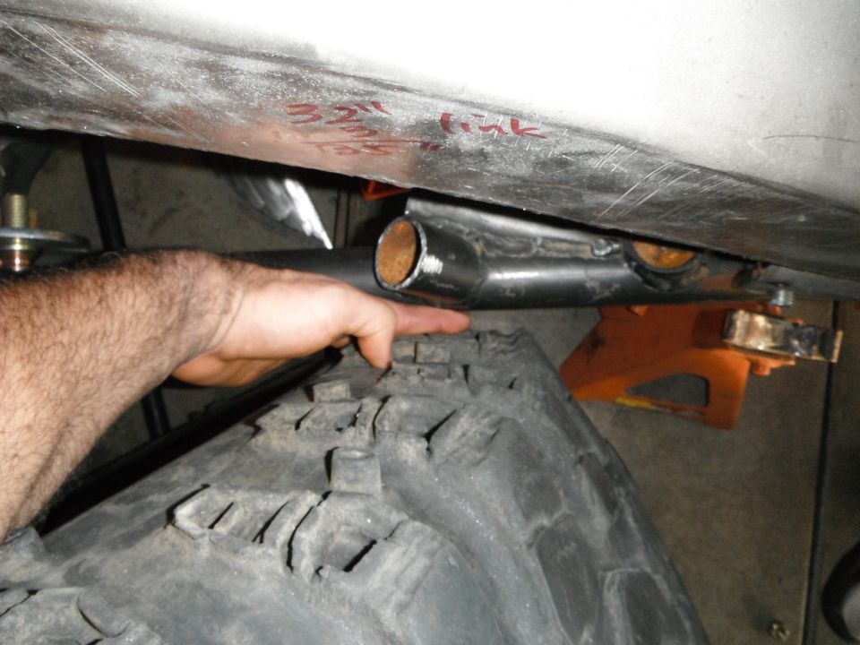 Undercarriage of a vehicle with a large tire, suspension linkage, and a hand pointing to the linkage. Red text reads "32" link"