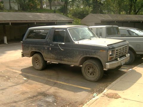 1982 Ford Bronco - A muddy dark blue Ford Bronco is parked in a lot next to another vehicle.