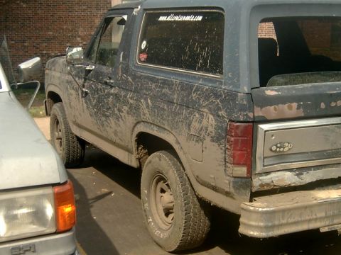 1982 Ford Bronco - Mud-splattered Ford Bronco, likely from the 1980s, parked beside another vehicle. A website URL is visible on the rear window.