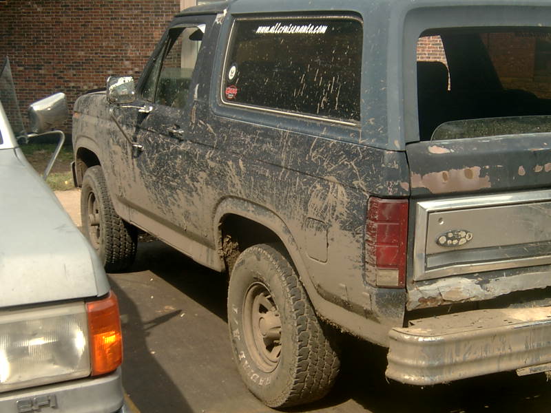 Mud-caked dark blue Ford Bronco rear quarter with open top and dented bumper