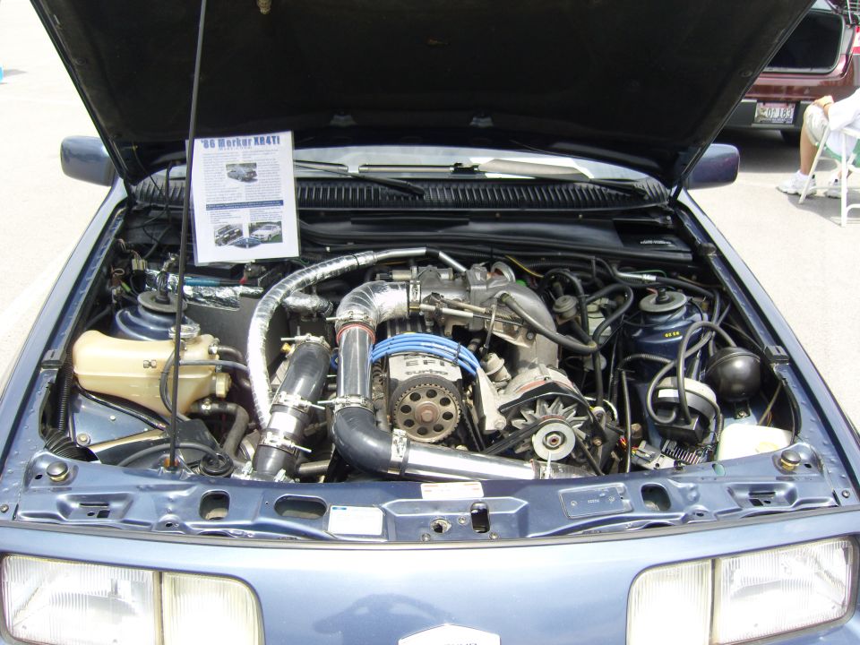 Engine bay of a blue 1986 Mercury XR4Ti showing a custom turbocharger setup and intercooler piping. A document is attached to the hood