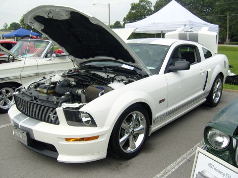 2007 Ford Mustang - A white 2007 Ford Mustang Shelby GT is displayed with its hood open, revealing the engine bay. Racing stripes adorn the hood and trunk.
