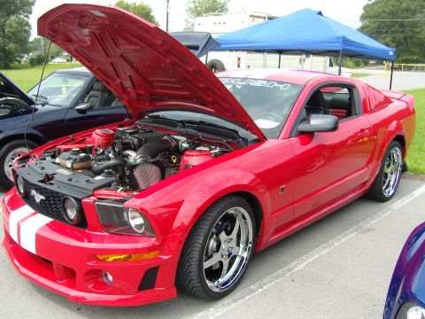 2007 Ford Mustang - A red Ford Mustang with racing stripes and an open hood displaying its supercharged engine is parked at an outdoor event.