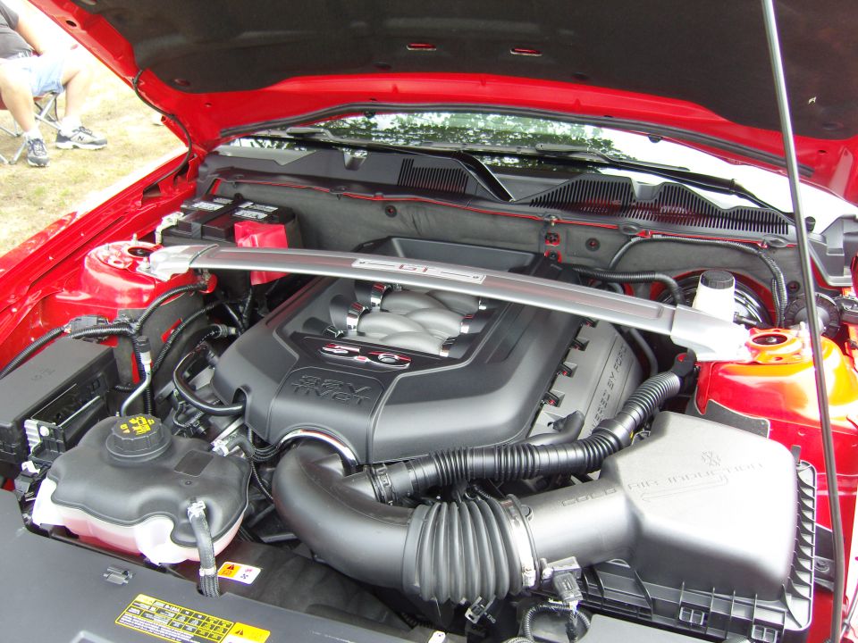 Engine bay of a red Ford Mustang, showing the 5.0 V8 engine cover and intake system