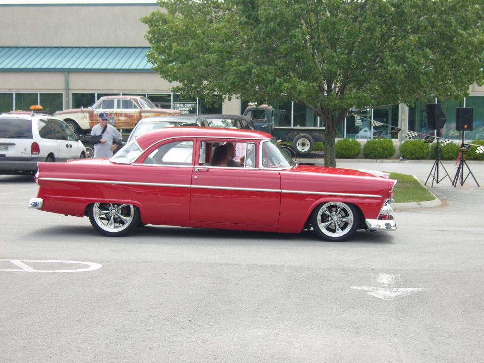 Red classic sedan with chrome trim and custom wheels parked outside a building. Another car and a van are visible behind it
