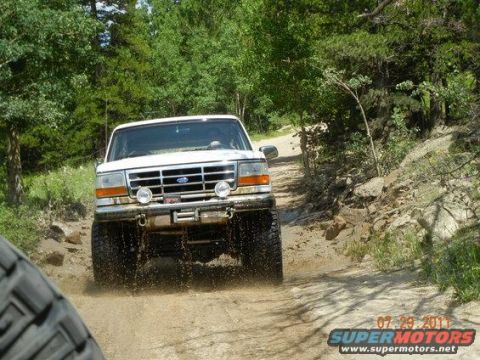 1995 Ford Bronco - A white 1990s Ford Bronco splashing water off its tires as it comes head-on up a rocky dirt trail through pine woods, lifted on mud tires w…