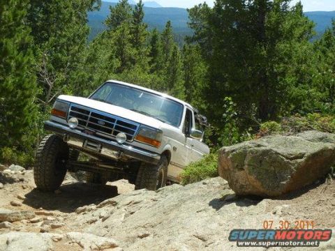 1995 Ford Bronco - A lifted white Ford Bronco with a chrome grille noses down a rocky high-country 4x4 trail lined with evergreens, a mountain ridge visible i…