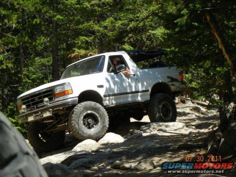 1995 Ford Bronco - Lifted white Ford Bronco with its hardtop removed crawling over a boulder-strewn trail in a pine forest, riding large mud-terrain tires on…