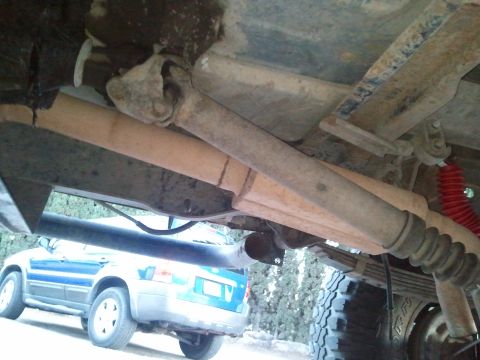 1989 Ford Bronco - Underbody view of a lifted 4x4 looking up at a surface-rusted two-piece driveshaft with U-joints, the frame rails, a leaf-spring pack and a…