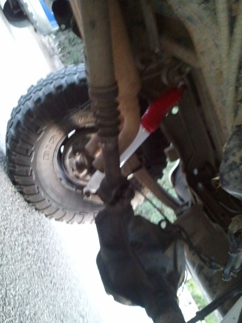 1989 Ford Bronco - Blurry underbody shot of a lifted 4x4 front suspension, showing rusty radius arms, spring seats and a red-booted shock absorber alongside a…