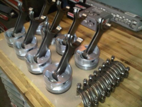 1979 Ford Bronco - Eight forged pistons with valve-relief dish tops stand assembled on their connecting rods, lined up on paper over a butcher-block workbench…