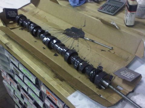 1979 Ford Bronco - A freshly unboxed V8 camshaft coated in black assembly lube resting in its long cardboard shipping carton on a workbench, with the distribu…