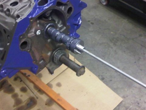 1979 Ford Bronco - A camshaft being slid into the front of a blue-painted V8 engine block on a stand, guided by a long threaded install rod threaded into the…